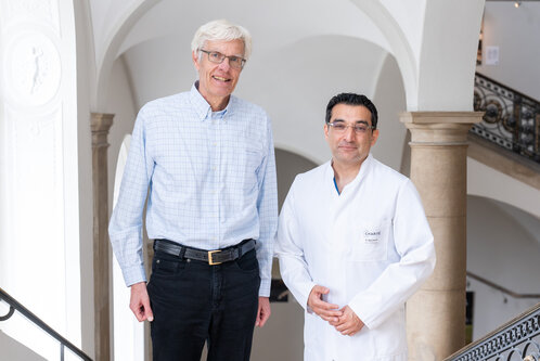 Zwei Männer stehen auf einer Treppe. Der Mann links trägt ein karierter Hemd und Jeans, der Mann rechts ist im weißen Arztkittel. Beide lächeln in die Kamera und stehen vor einem architektonisch ansprechenden Hintergrund.