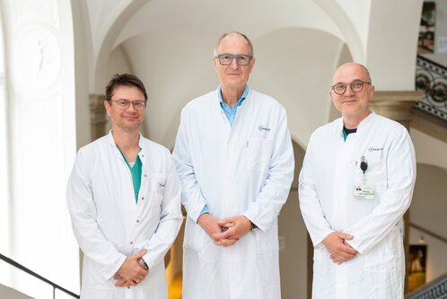 Drei medizinische Fachkräfte in weißen Kitteln stehen lächelnd in einem hellen Innenraum. Sie wirken freundlich und professionell, bereit, ihre Patienten zu unterstützen. Im Hintergrund sind architektonische Details sichtbar.