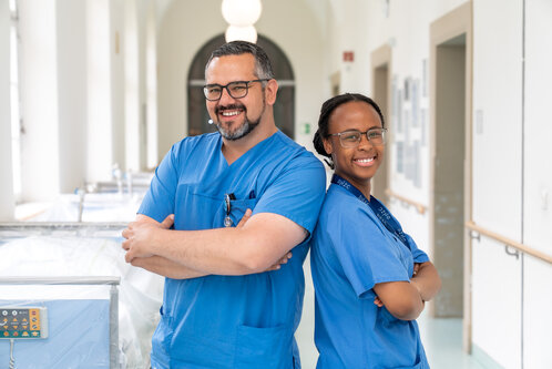 Zwei medizinische Fachkräfte in blauen Arbeitsuniformen stehen Rücken an Rücken in einem Krankenhausflur. Sie lächeln und strahlen Selbstvertrauen aus, während im Hintergrund mehrere Krankenhausbetten sichtbar sind.
