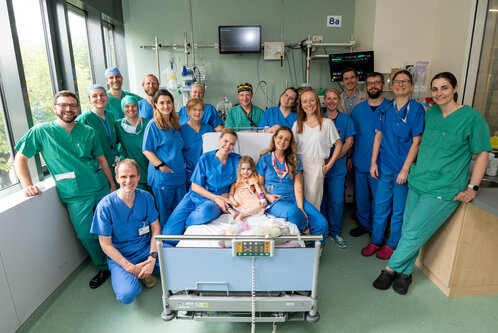 Eine Gruppe von über 20 medizinischen Fachkräften, darunter Ärzte und Pflegekräfte, posiert lächelnd um ein Krankenbett, in dem ein Kind mit einer Umarmung sitzt. Alle sind in grünen und blauen Krankenhauskleidern gekleidet. Die Szene vermittelt eine Atmosphäre der Unterstützung und Fürsorge.