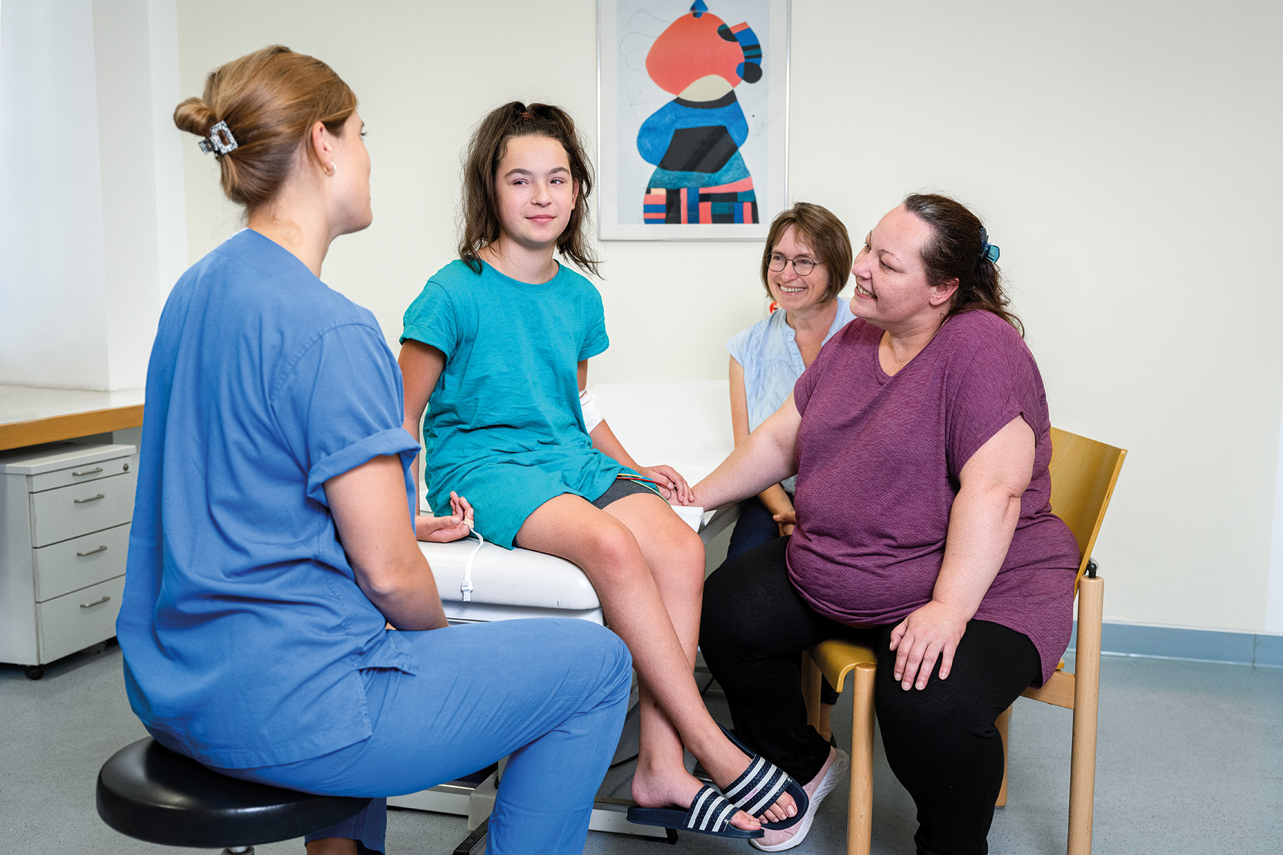Eine junge Patientin sitzt auf einem Behandlungstisch und spricht mit zwei Frauen in blauer Arbeitskleidung. Im Hintergrund sind zwei weitere Frauen, die aufmerksam zuhören. Alle wirken freundlich und kommunikativ, was eine unterstützende Atmosphäre schafft.