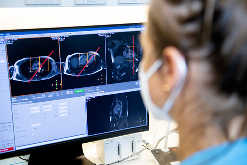 Eine Person betrachtet einen Computerbildschirm mit medizinischen Bildgebungen. Auf dem Bildschirm sind mehrere radiologische Bilder zu sehen, die vermutlich von einer Diagnose stammen. Die Person trägt eine Maske und hört aufmerksam zu.