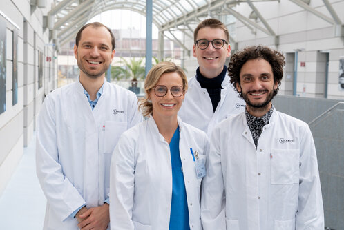 Vier Personen in weißen Laborkitteln lächeln in einem modernen, hellen Innenraum. Sie stehen in einer Reihe, jeder mit unterschiedlichen Frisuren und Gesichtsausdrücken. Im Hintergrund sind Pflanzen und eine großen Fensterfront sichtbar.