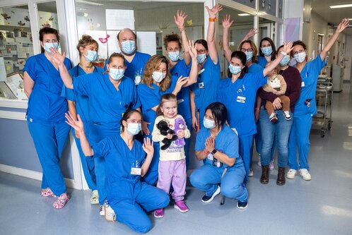 Eine Gruppe von elf Pflegekräften in blauen Uniformen und medizinischen Masken steht in einem Krankenhausflur. In der Mitte eines kleinen Mädchens, das Plüschtiere hält. Alle lächeln und heben die Hände in einer fröhlichen Geste.