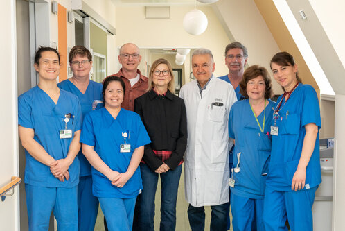Eine Gruppe von neun Personen steht in einem Krankenhausflur. Sie tragen blaue Krankenhauskleidung. In der Mitte steht ein Arzt in einem weißen Kittel, umgeben von sechs Mitarbeitern und zwei Besuchern. Alle lächeln und schauen in die Kamera.