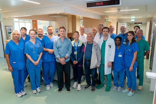 Eine Gruppe von medizinischem Personal in blauer Arbeitskleidung steht in einem Krankenhausflur. Sie lächeln und posieren gemeinsam für ein Foto. Im Vordergrund sind auch einige Besucher, die eng mit dem medizinischen Team verbunden sind.