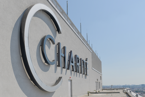 Das Bild zeigt das große, klare Logo der Charité auf einem modernen Gebäude. Der Schriftzug "Charité" ist prominent und basiert auf einem hellen Hintergrund, während der Himmel blau und klar ist. Es vermittelt den Eindruck eines bedeutenden Gesundheitseinrichtungsortes.
