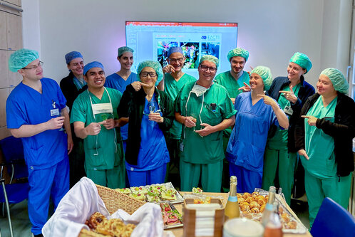 Eine Gruppe von Gesundheitsfachkräften in blauer und grüner Arbeitskleidung steht zusammen und lächelt. Sie halten Getränkeflaschen und befinden sich in einem Raum mit einem Tisch voller Snacks und Leckereien. Im Hintergrund ist ein Bildschirm sichtbar.
