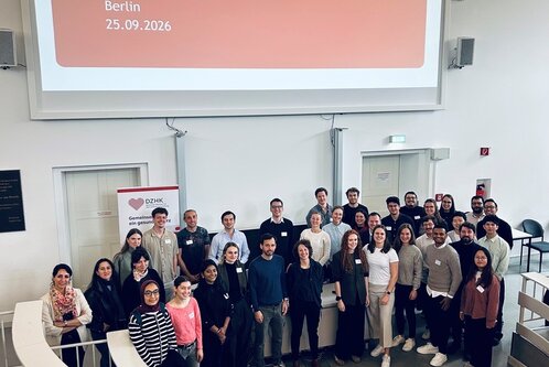 Eine Gruppe von Teilnehmern steht in einem modernen Konferenzraum vor einer Präsentationswand mit der Aufschrift "Young-DZHK Cardiovascular Research Summit 2026, Berlin, 25.09.2026". Die Anwesenden sind vielfältig und tragen unterschiedliche Kleidung, während sie lächeln und in die Kamera schauen.