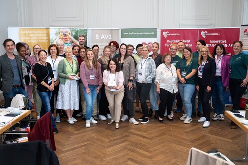 Eine Gruppe von etwa 30 Personen steht in einem Raum, lächelt und posiert für ein Gruppenfoto. Im Hintergrund sind Werbebanner sichtbar. Die Personen tragen unterschiedliche Kleidung und scheinen an einer Veranstaltung teilzunehmen, die gemeinschaftlich und freundlich wirkt.